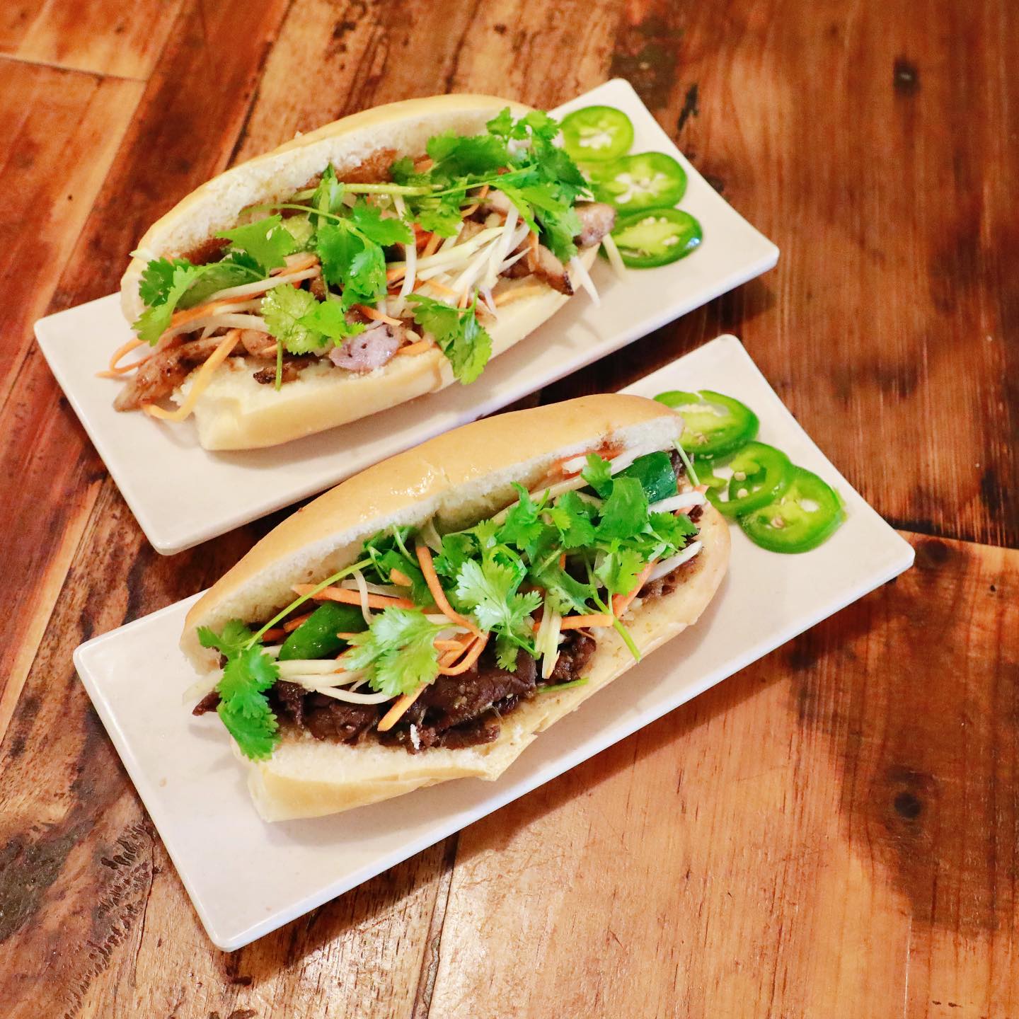 Two authentic Vietnamese bánh mì sandwiches with grilled pork and beef, fresh cilantro, pickled vegetables, jalapeños on white plates at La Cà restaurant in Tacoma