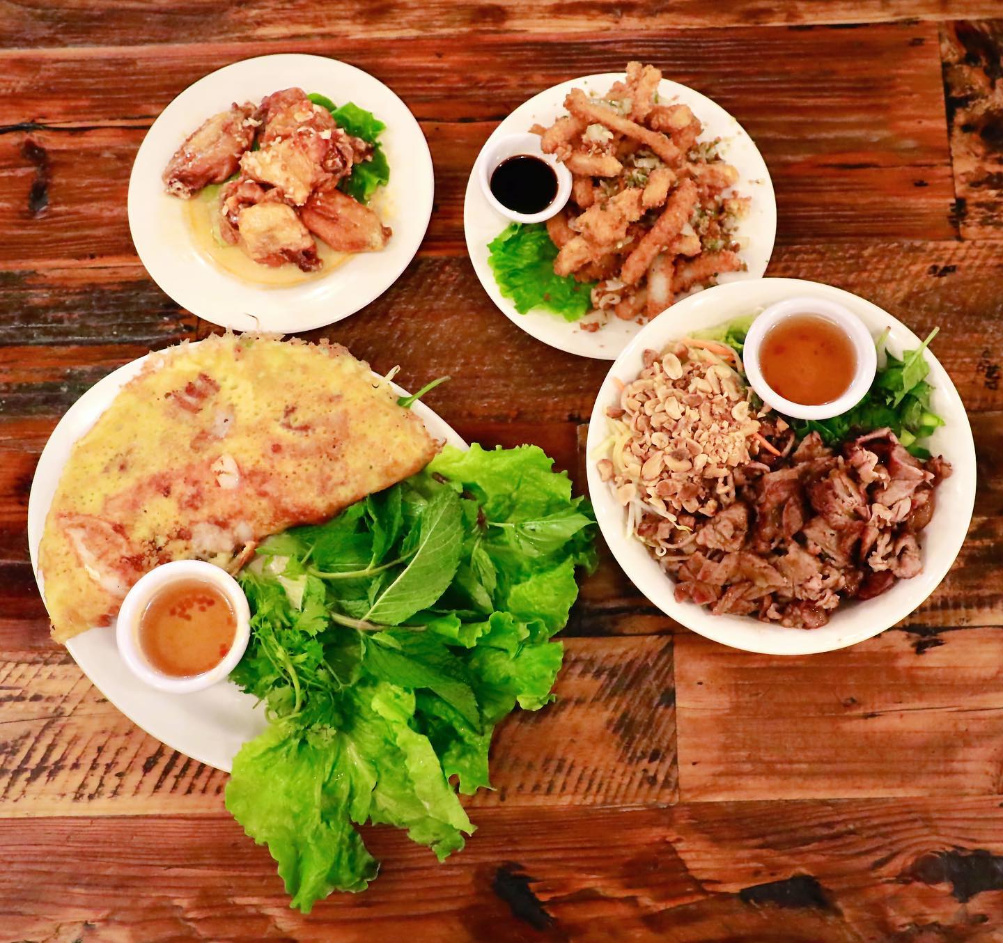 Overhead view of authentic Vietnamese food spread at La Cà 38th featuring bánh xèo crispy crepe, salt and pepper squid, fried calamari, sliced beef with peanuts and noodles, fresh herbs, and traditional dipping sauces on rustic wood table in Tacoma